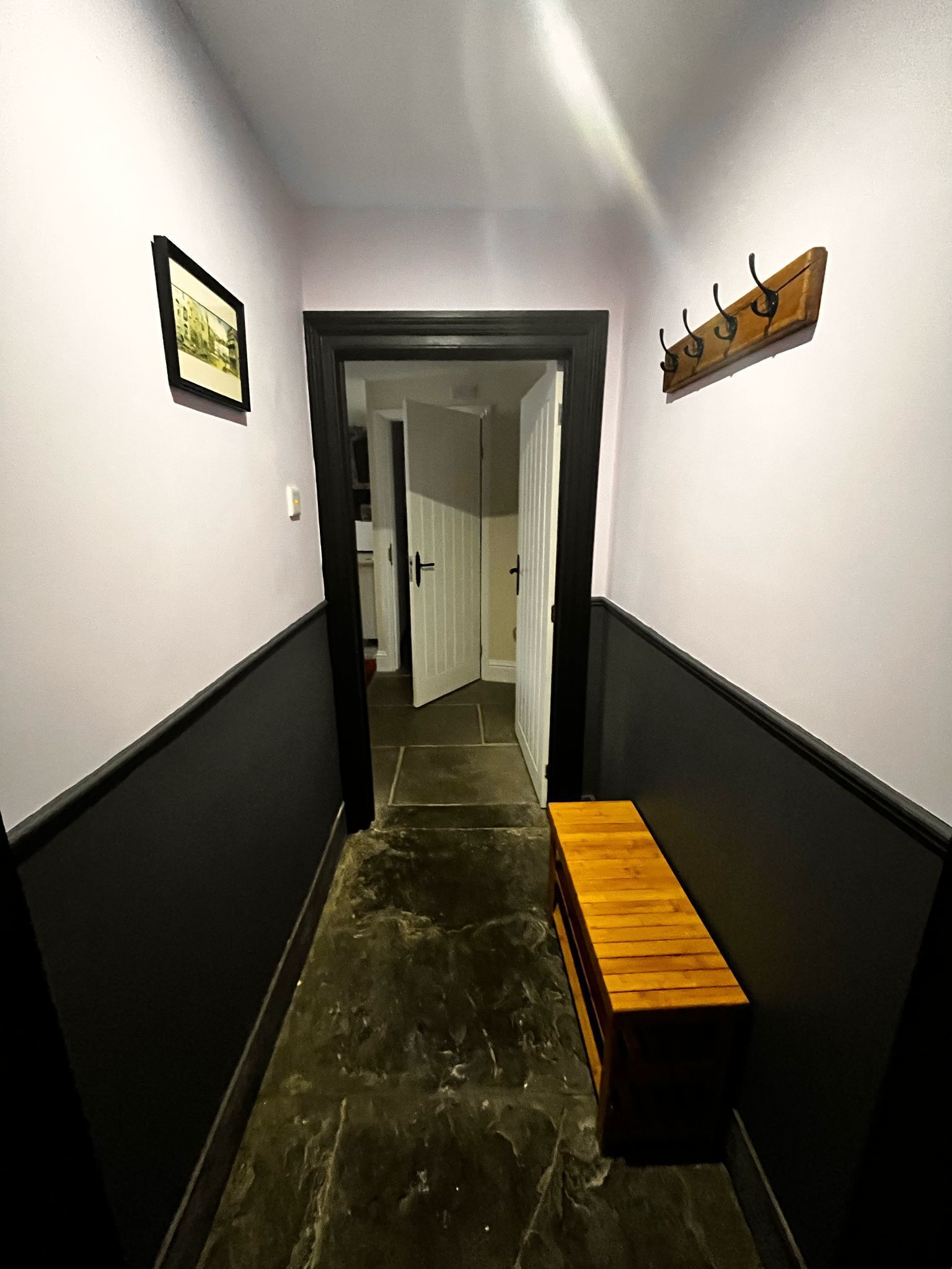 Un pasillo estrecho con un banco de madera, una puerta al final y un cuadro en la pared.