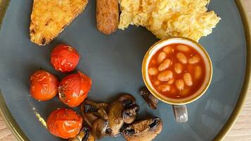 Um prato com uma fatia de peixe frito, linguiça, ovos mexidos, tomate assado, cogumelos e uma chávena de feijão cozido.