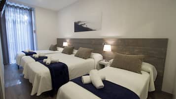 A hotel room with three beds, each with a white bedspread and a dark blue blanket. There are two bedside lamps on each bed, and a painting on the wall above the beds.