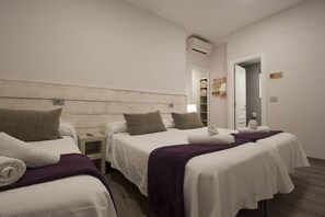 Une chambre d’hôtel avec deux lits, chacun étant équipé de draps blancs et d’un plaid violet. On y trouve une petite table de chevet avec une lampe et un téléphone. La chambre est dotée d’une tête de lit en bois et d’un mur blanc sur lequel est installé un petit appareil de climatisation.