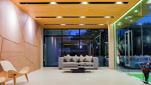 A modern lobby with a wooden ceiling, a grey sofa with cushions, two wooden chairs, and a glass wall with a view of the outside.