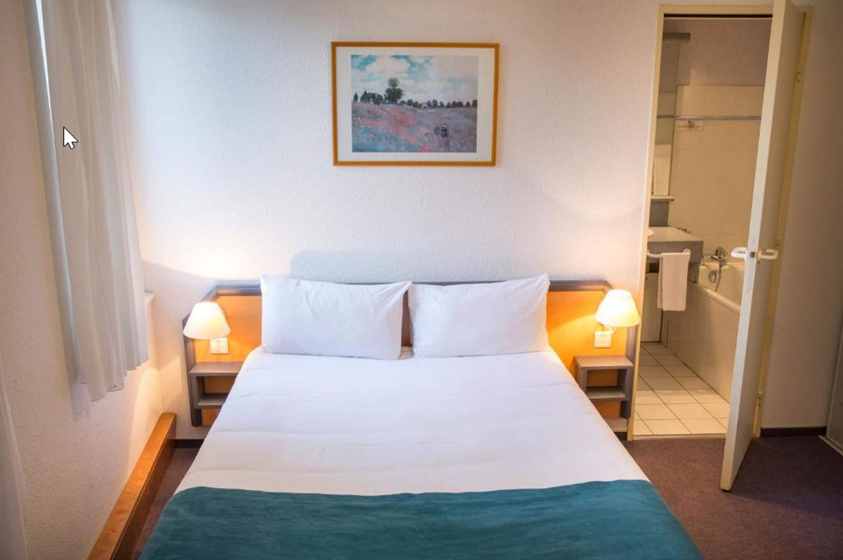 Une chambre d’hôtel avec un lit, deux lampes de chevet, un tableau au mur et une salle de bain visible à travers une porte ouverte.