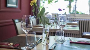 Una mesa de comedor con copas de vino vacías, un jarrón con una sola flor amarilla y una vista despejada del interior del restaurante.