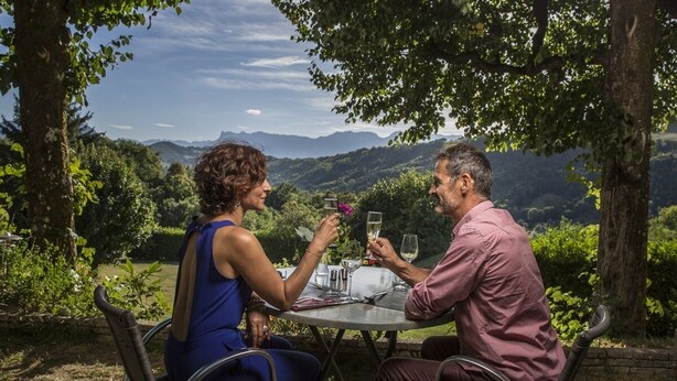 Dos personas sentadas en una mesa al aire libre, con una vista panorámica de colinas al fondo.