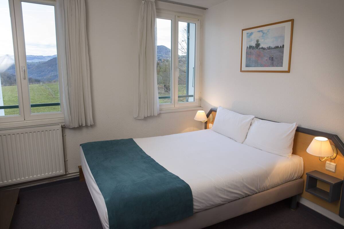 A hotel room with a bed, two windows, a painting, and a nightstand with a lamp.