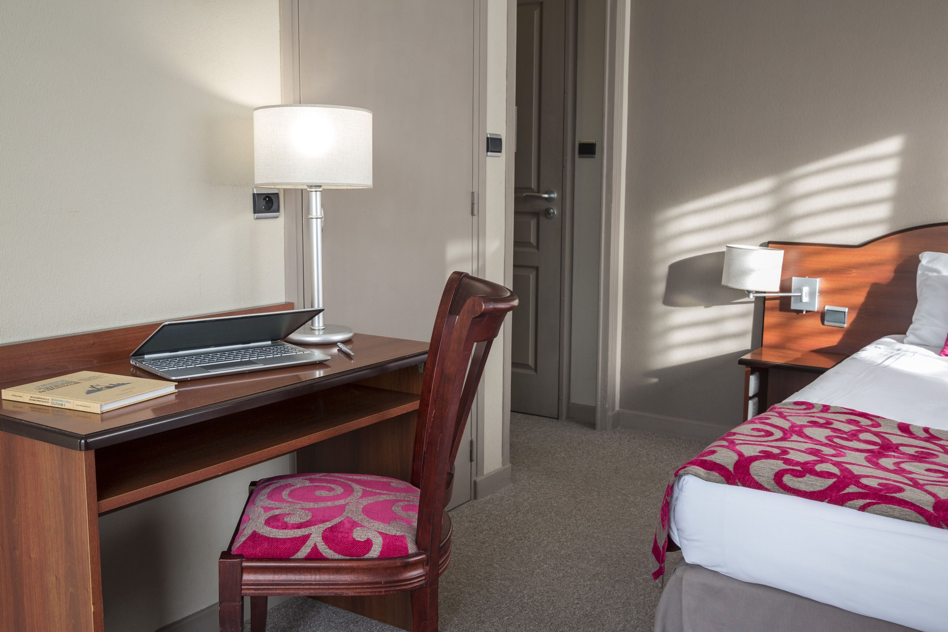 A hotel room with a wooden desk, a laptop, a chair, a bed with a patterned bedspread, and a nightstand with a lamp.