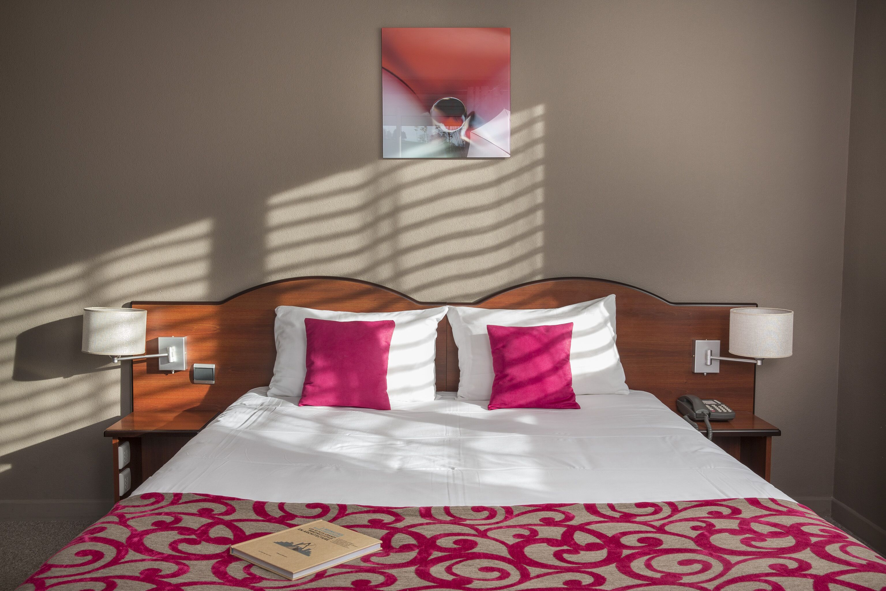 A hotel room with a neatly made bed, two pillows, a bedside lamp, a telephone, and a framed picture on the wall.