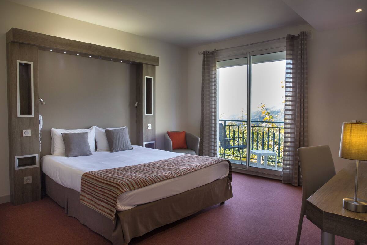 A hotel room with a large bed, a desk, a chair, a balcony with a view, and a window with curtains.