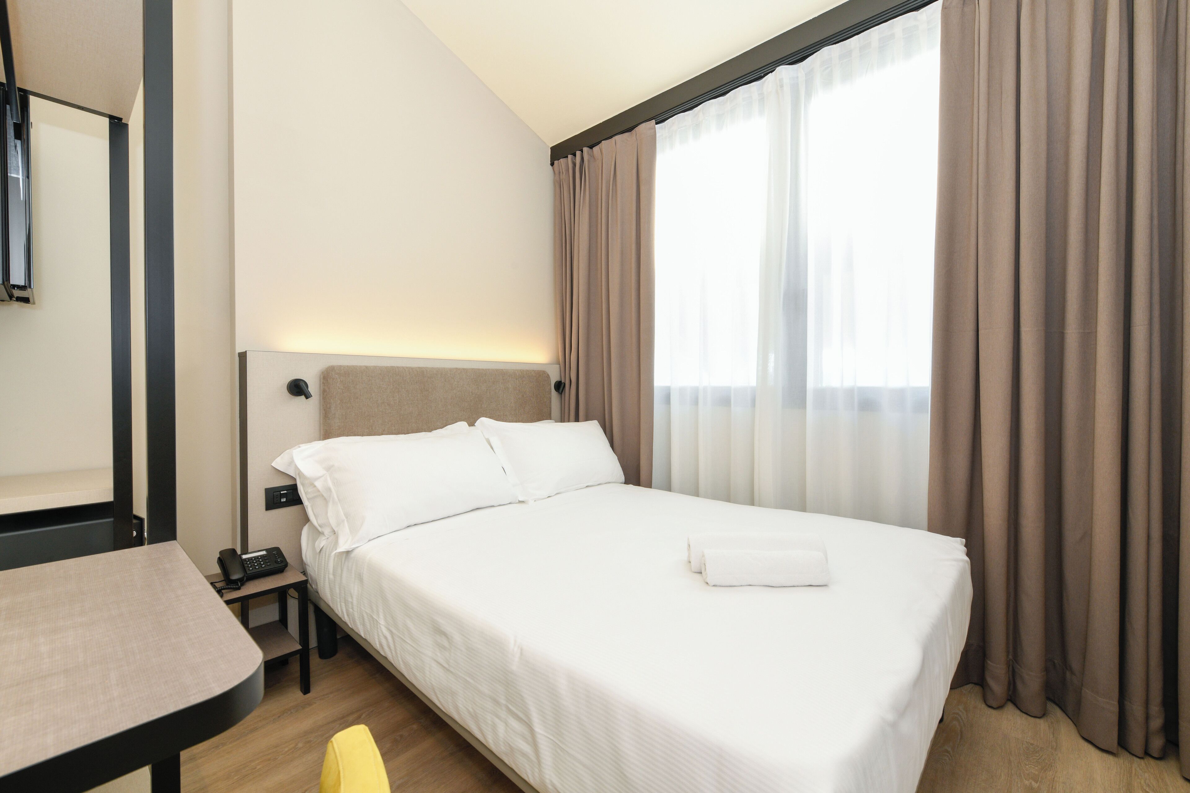 Ein Hotelzimmer mit einem Bett, einem Nachttisch mit Telefon, einem kleinen Tisch und einem Fenster mit Vorhängen.