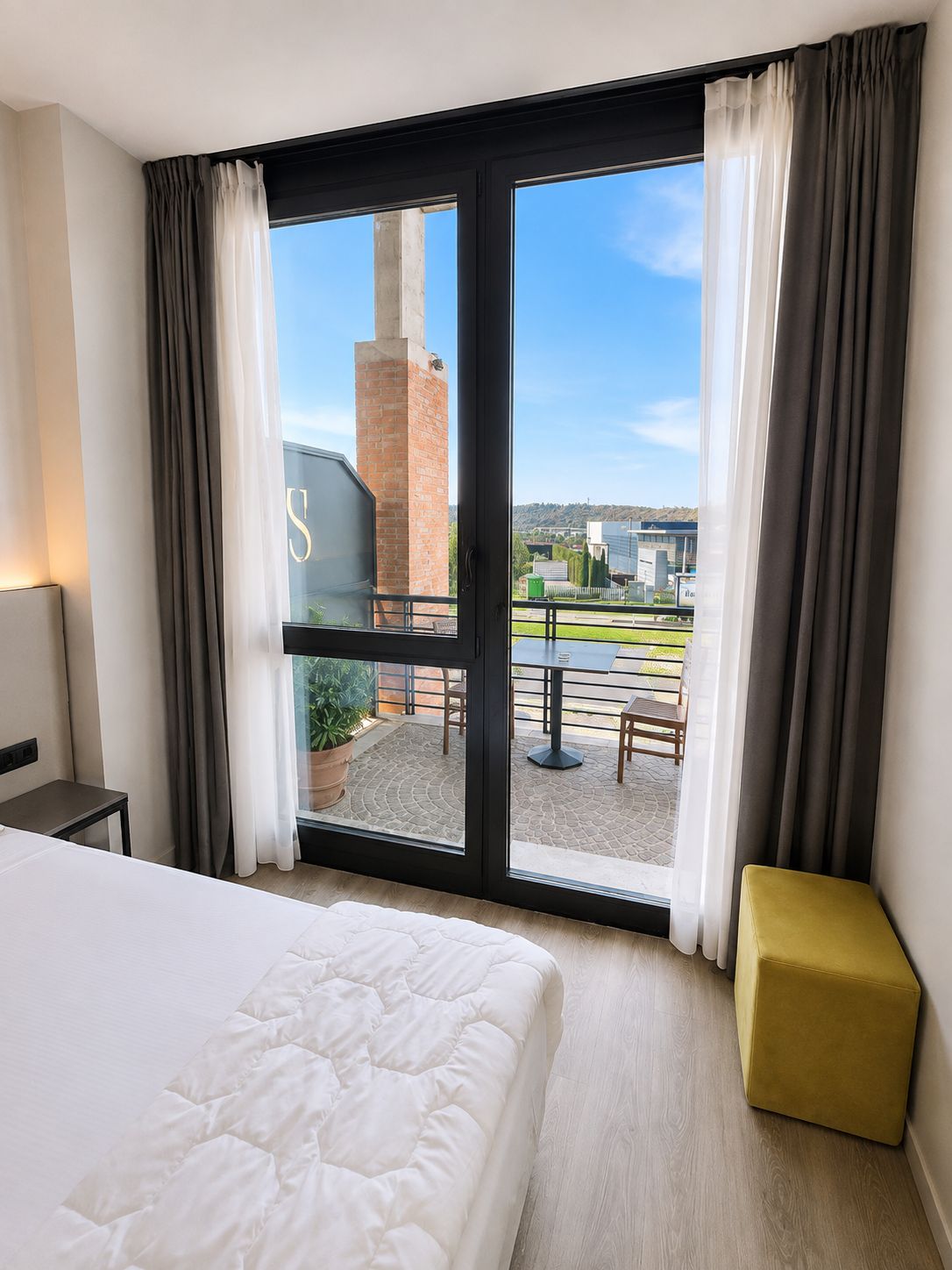 A hotel room with a bed, a small table, a bench, and a large window with curtains.