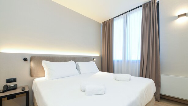 A neatly made bed with white linens and two towels, a bedside table with a phone and remote, and a window with curtains. - Best Western Hotel Garda (Affi)