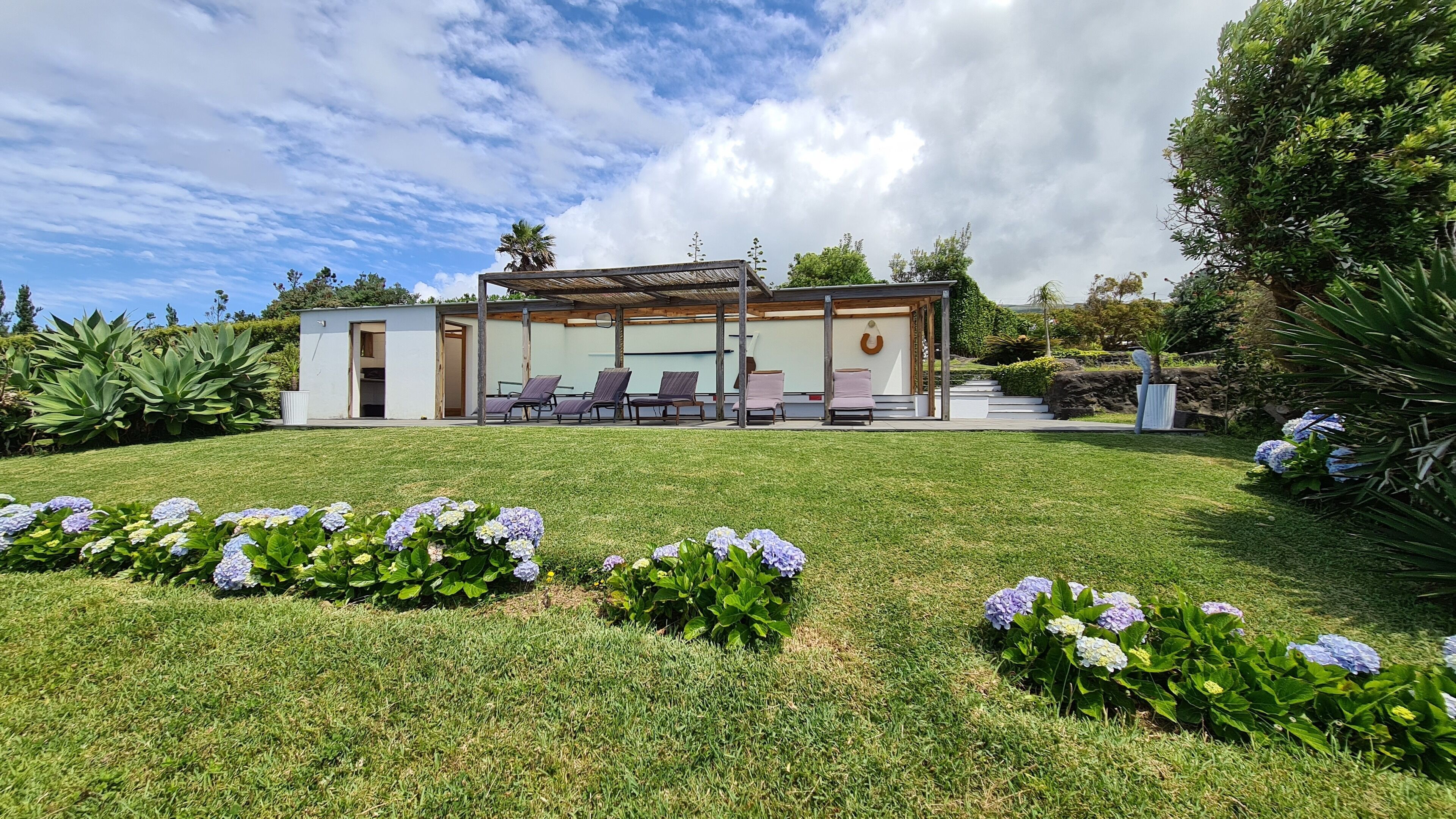Una casa moderna con un área de estar cubierta al aire libre, un pequeño jardín con flores de hortensia y un cielo azul despejado.