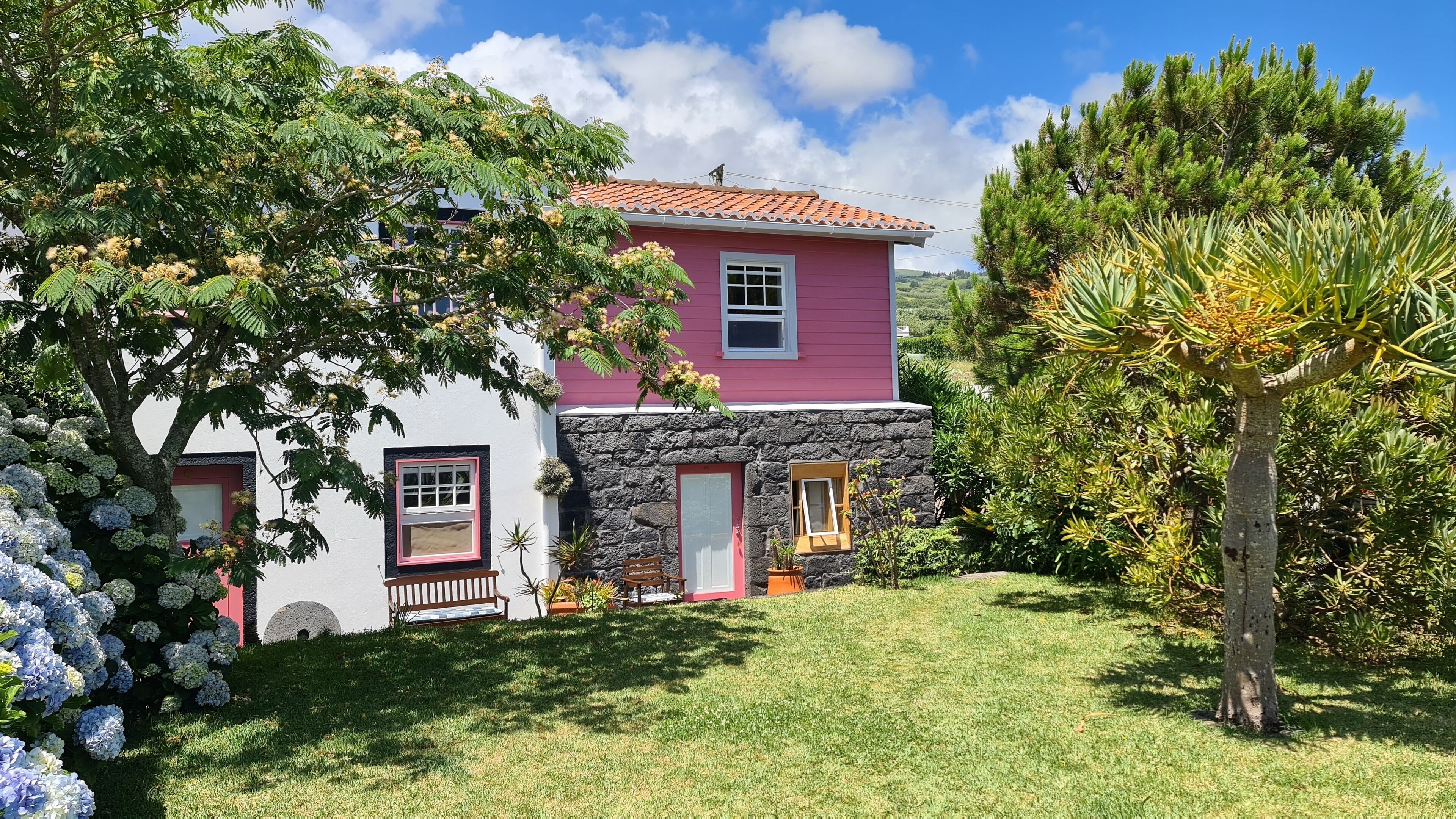 Una casa de dos pisos con fachada de piedra y exterior rosa, y un jardín bien cuidado con hortensias y una palmera.