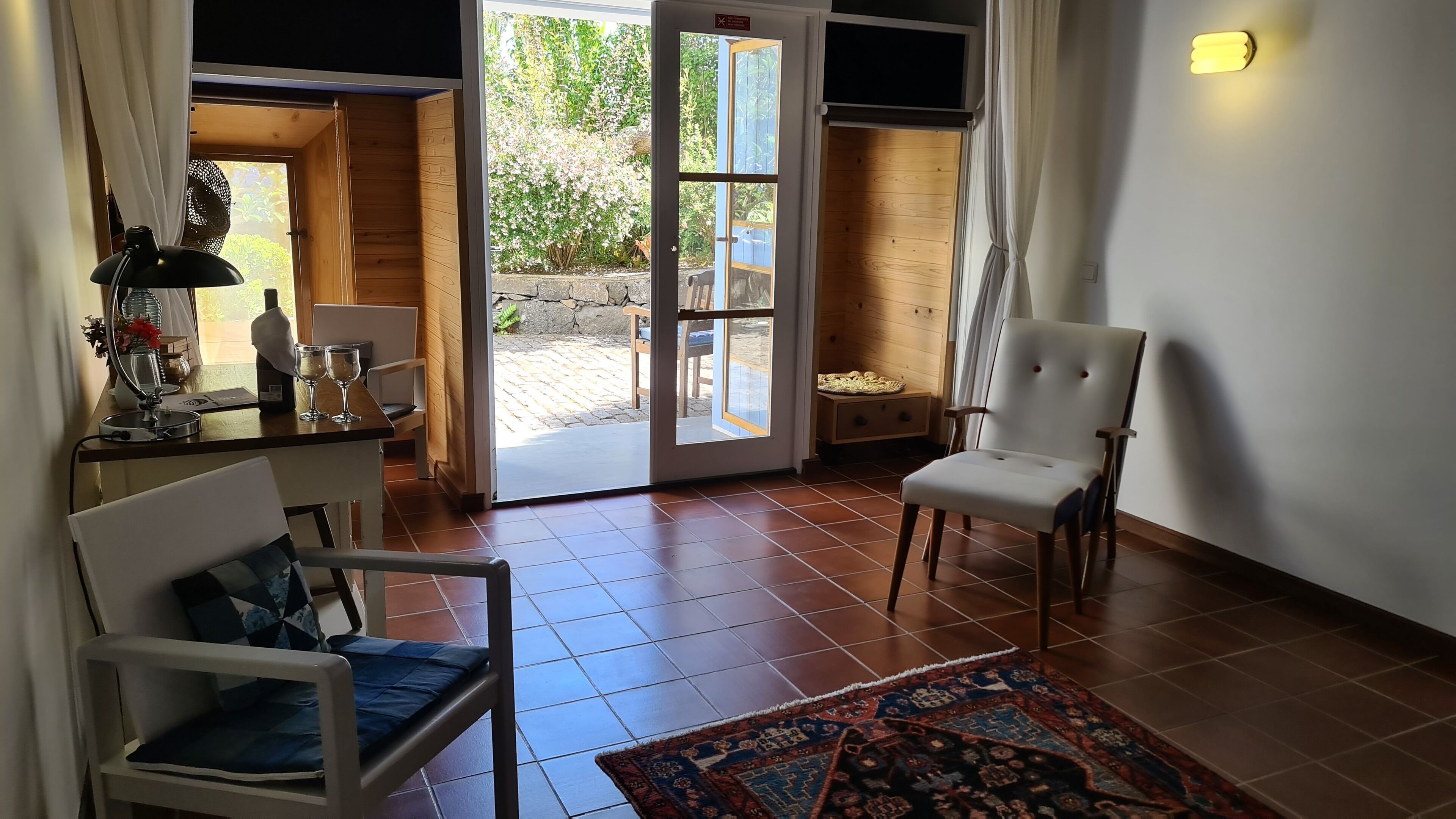 Una stanza con arredamento in legno, una sedia bianca, un tavolo con specchio e bicchieri, e vista su una zona esterna con panchina in legno e vegetazione.