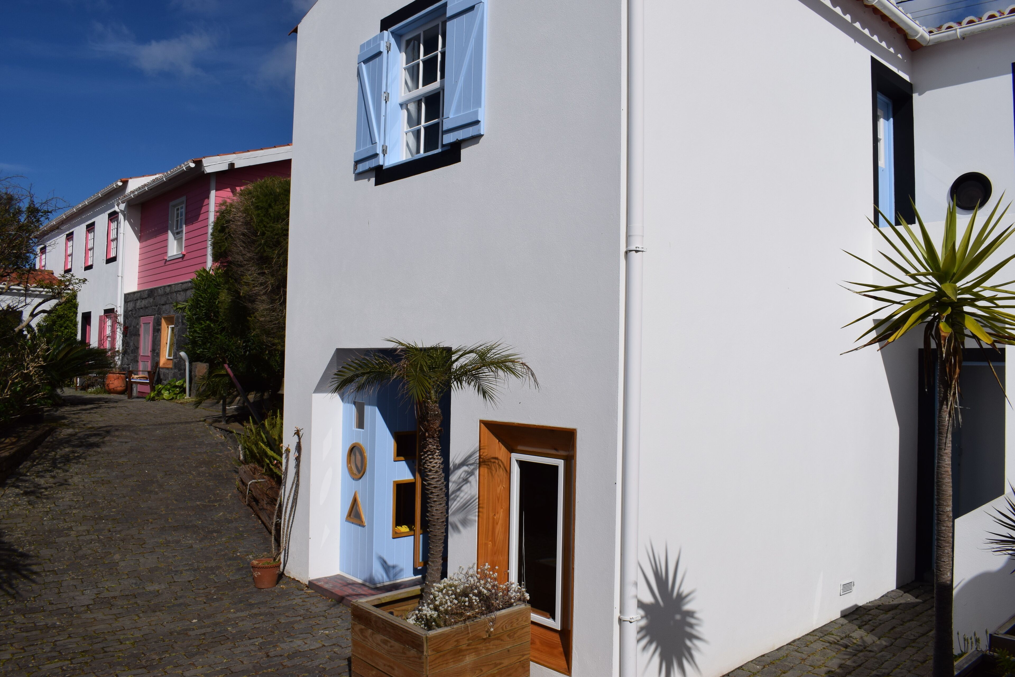 A narrow cobblestone street flanked by colorful buildings with distinct architectural features, including a white building with a wooden door and a blue window, and a pink building with a blue window.