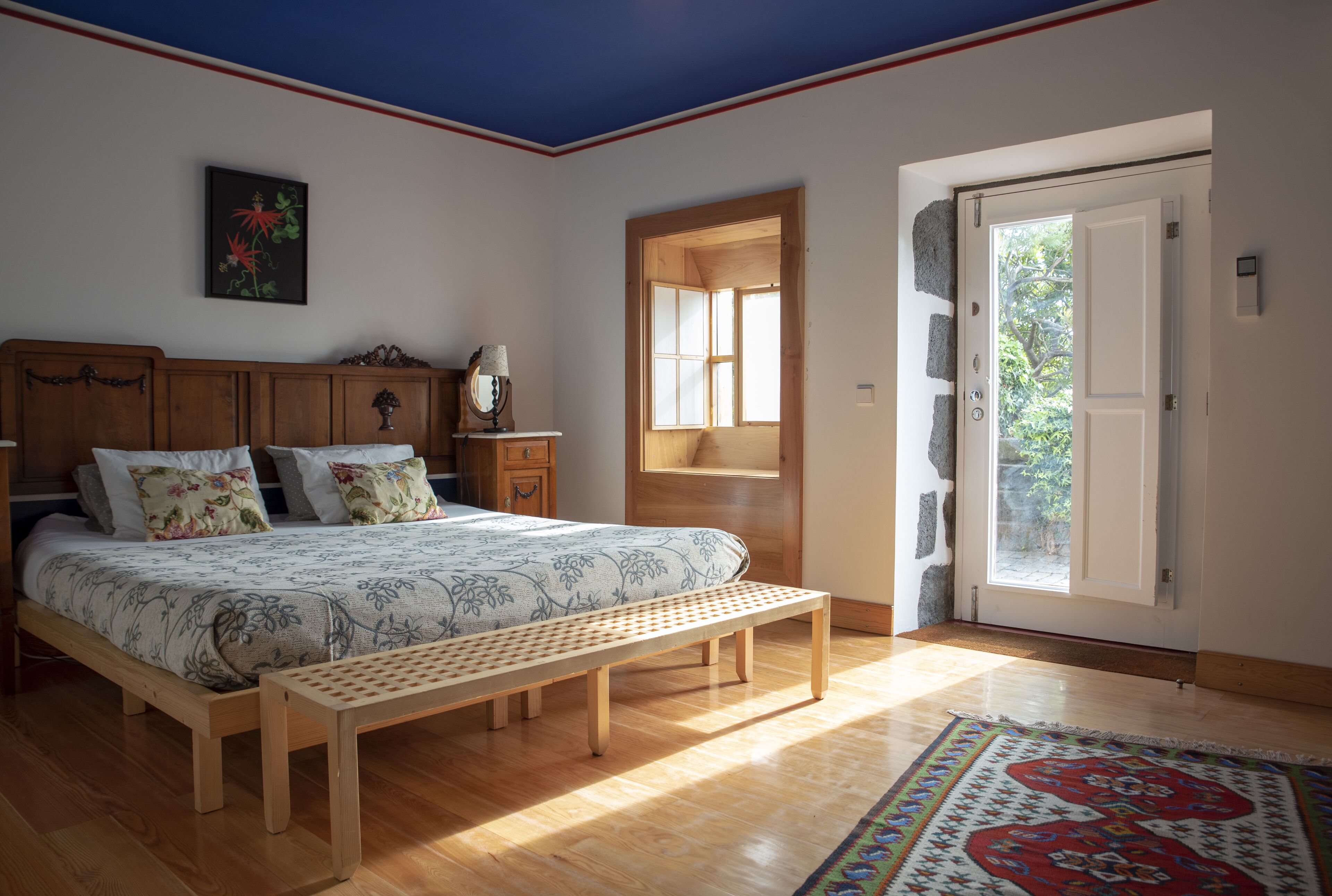 Un dormitorio con una cama de madera, un banco, una mesita de noche con lámpara, una ventana y una puerta que da al exterior.