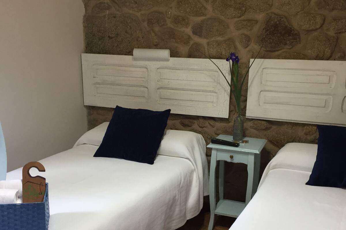 Quarto com duas camas, roupa de cama branca, parede de pedra e uma pequena mesa com um vaso.