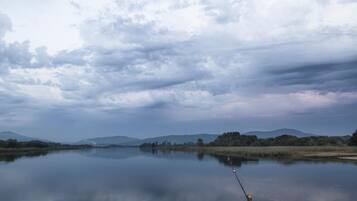 A calm lake with a fishing rod in the foreground, surrounded by hills and trees under a cloudy sky.
