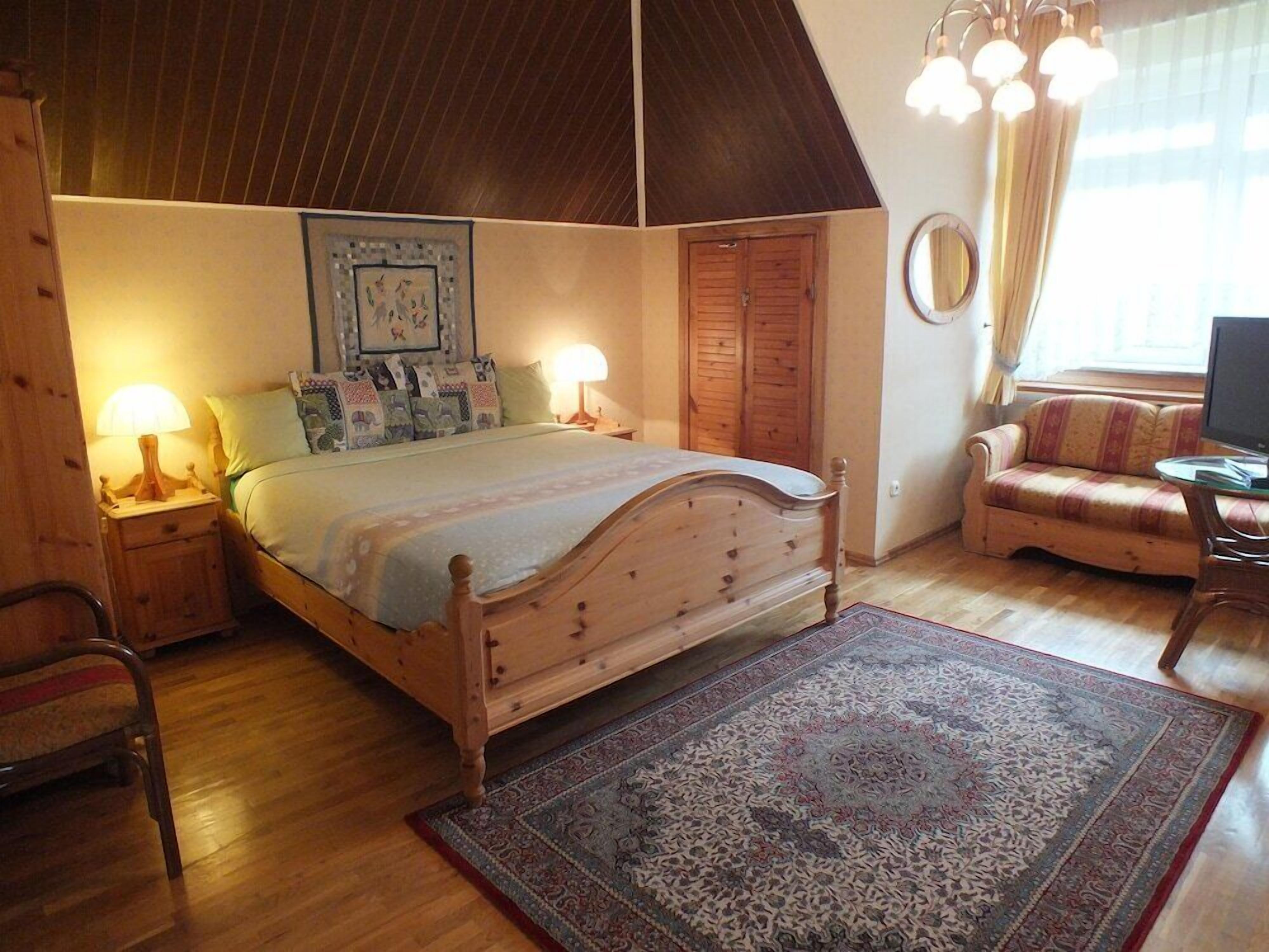 A bedroom with a wooden bed, two bedside tables with lamps, a sofa, a television, and a patterned rug.