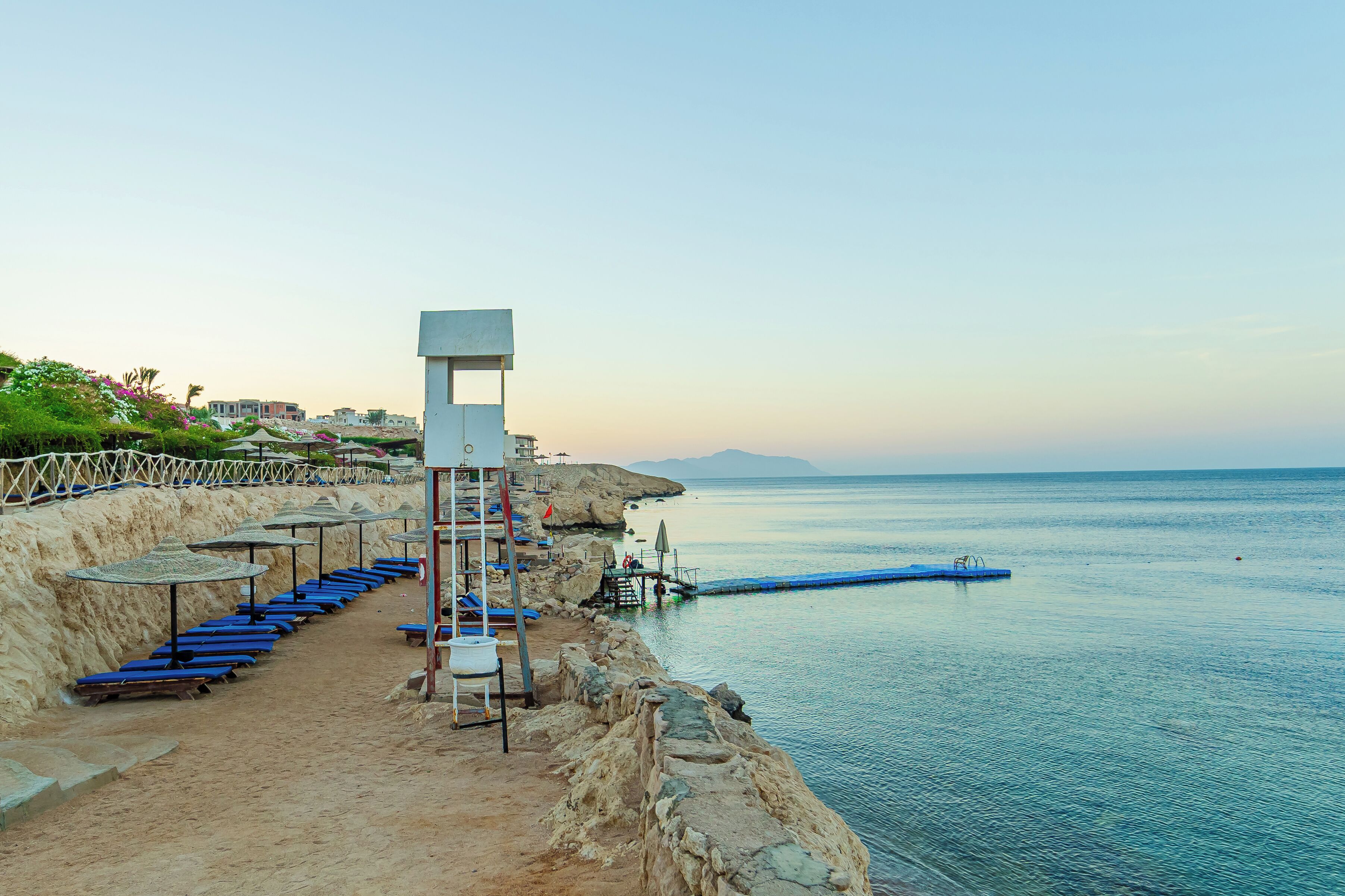 A coastal walkway with blue lounge chairs, a lifeguard tower, and a rocky shoreline.