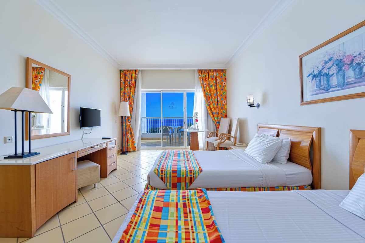 A hotel room with a bed, a desk with a TV, a chair, a balcony with a view, and a painting on the wall.