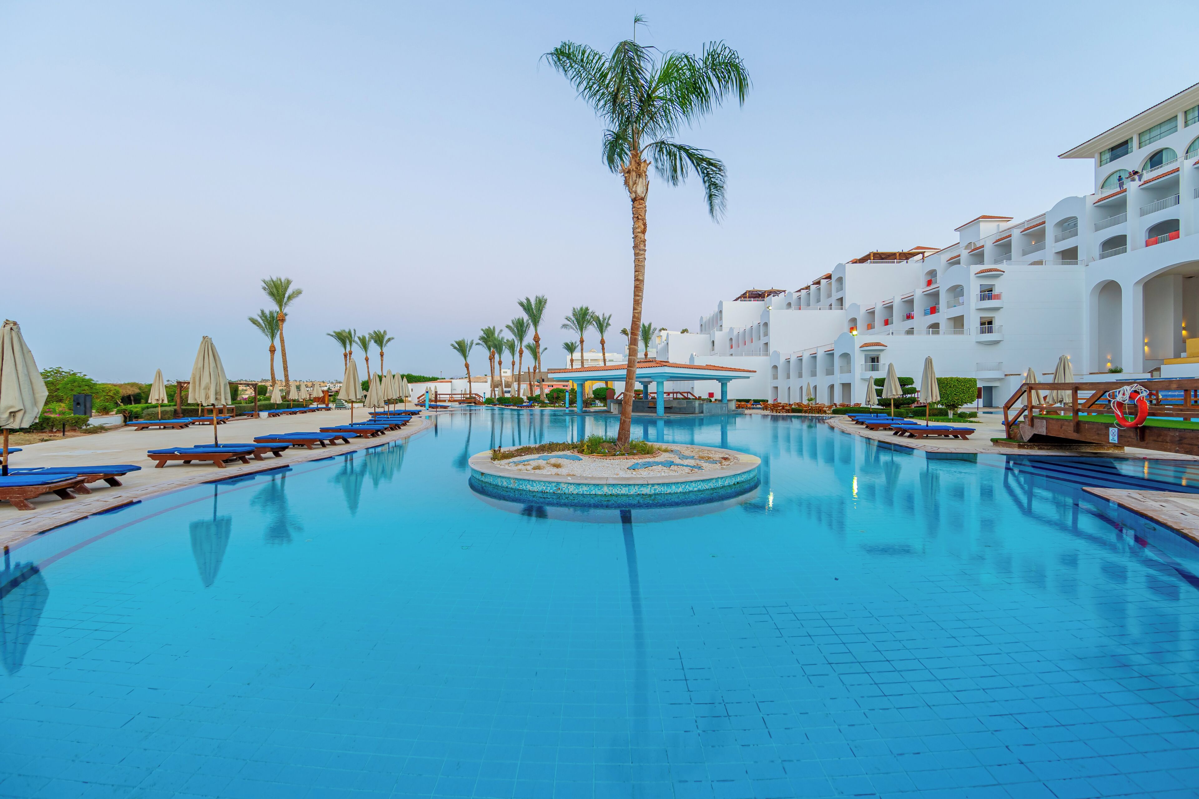 Ein Schwimmbecken mit Liegestühlen, Palmen und einem Hotelgebäude mit Balkonen.