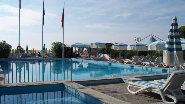 A swimming pool with lounge chairs and umbrellas, surrounded by flags and a fence. - Monaco Quisisana (Jesolo)