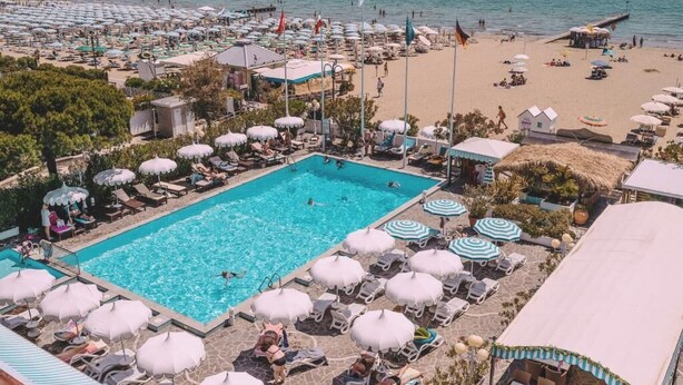 A resort with a swimming pool surrounded by sun loungers and umbrellas, a beach with umbrellas and a pier, and a clear view of the sea. - Monaco Quisisana (Jesolo)