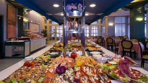 A buffet with seafood and salads spread out on a long table, with a central decorative column and a seating area in the background. - Monaco Quisisana (Jesolo)