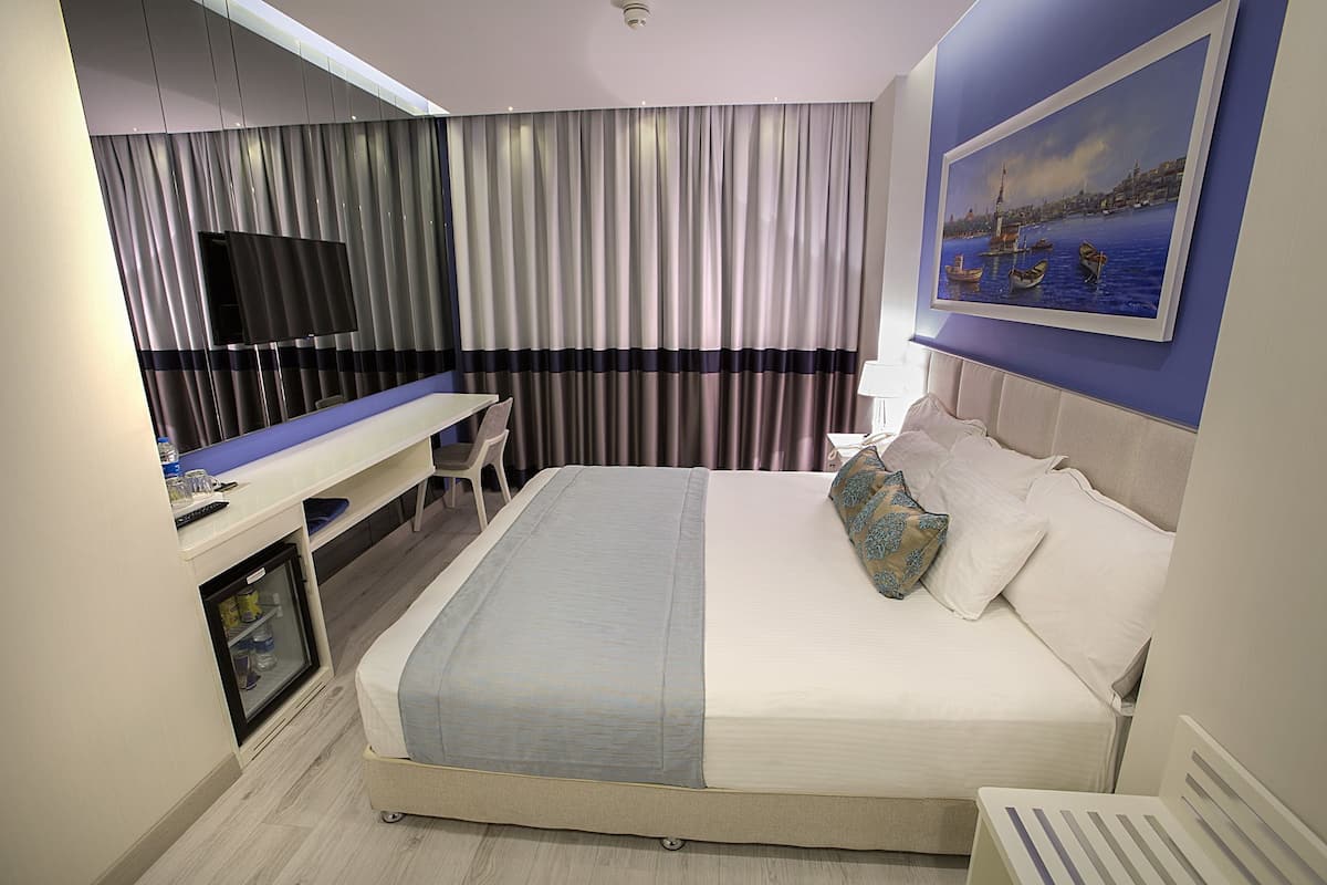 A modern hotel room with a bed, a desk, a television, and a painting on the wall.