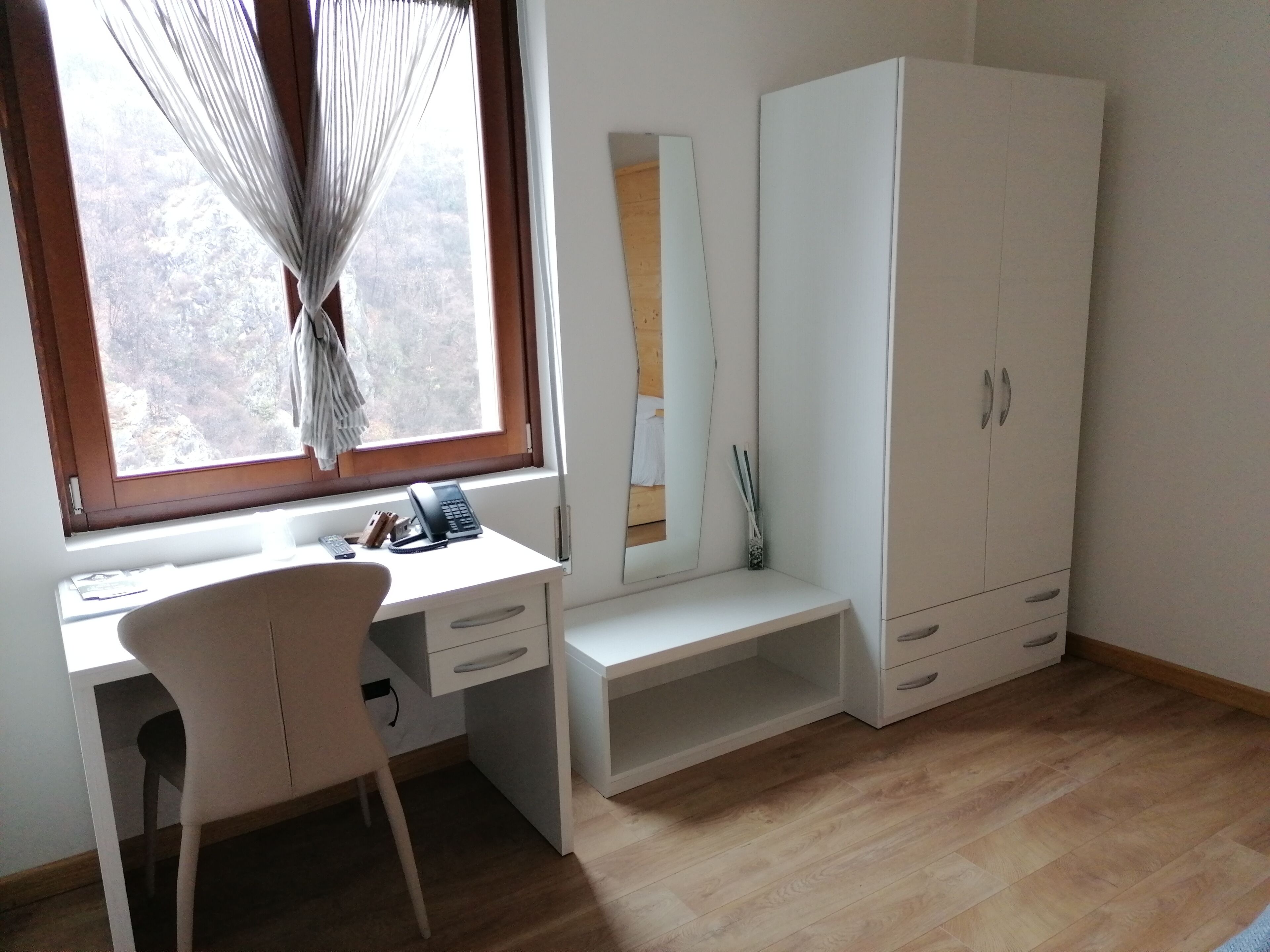 A room with a desk, chair, wardrobe, and a window with curtains.