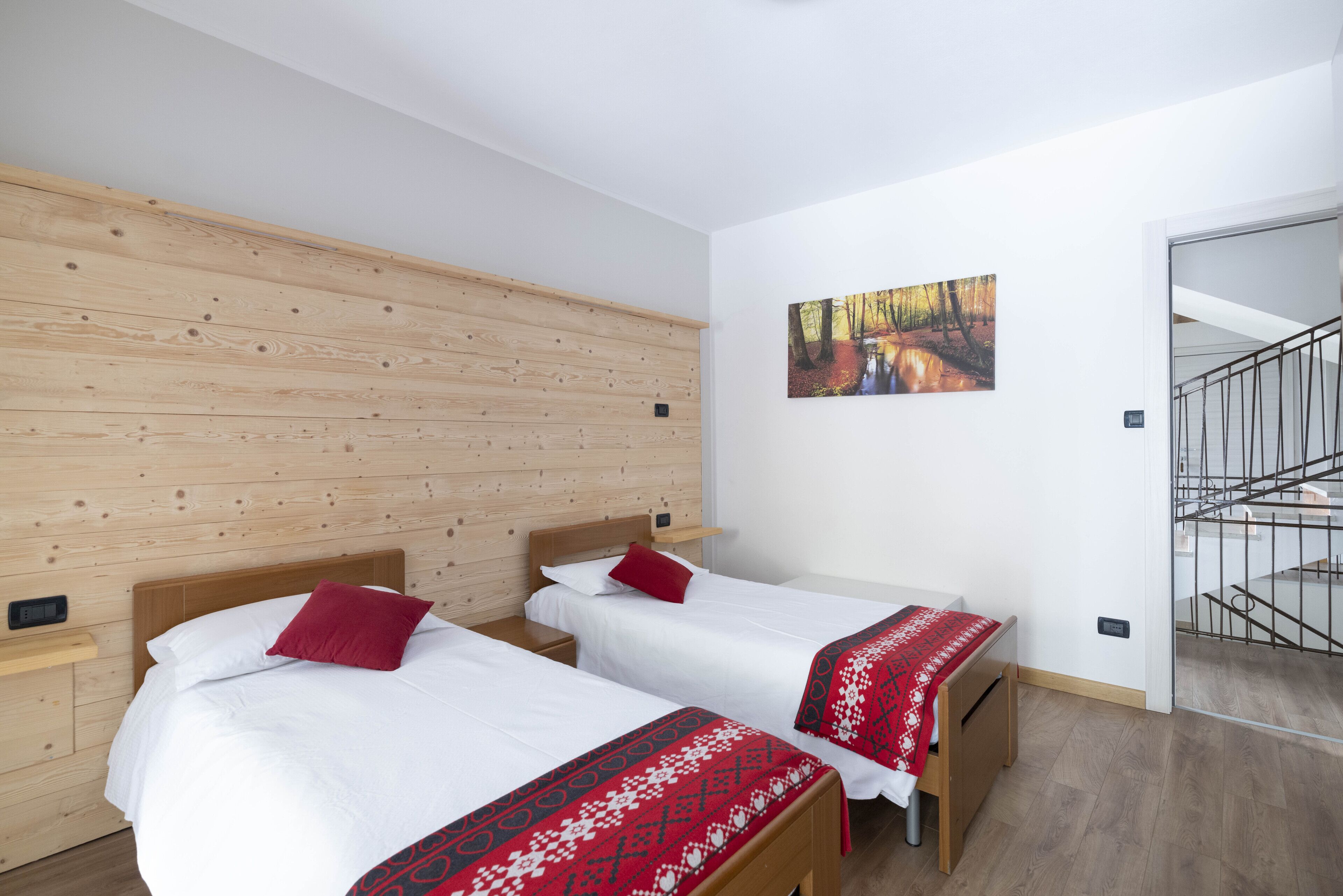 A hotel room with two beds, wooden panel walls, a painting of a forest, and a staircase.