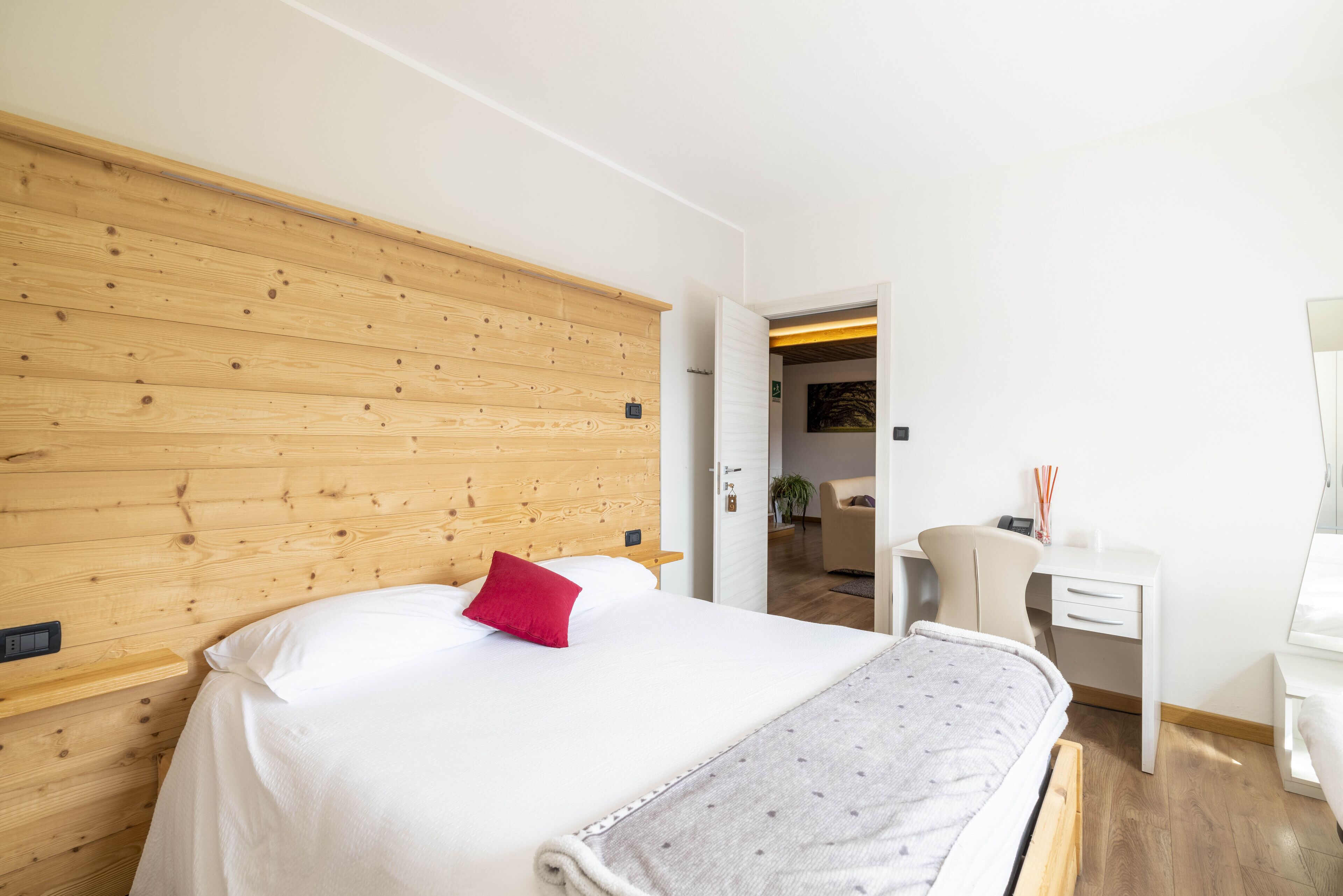A bedroom with a wooden headboard, a bed with white bedding and a red pillow, a desk with a chair, and a small bedside table.