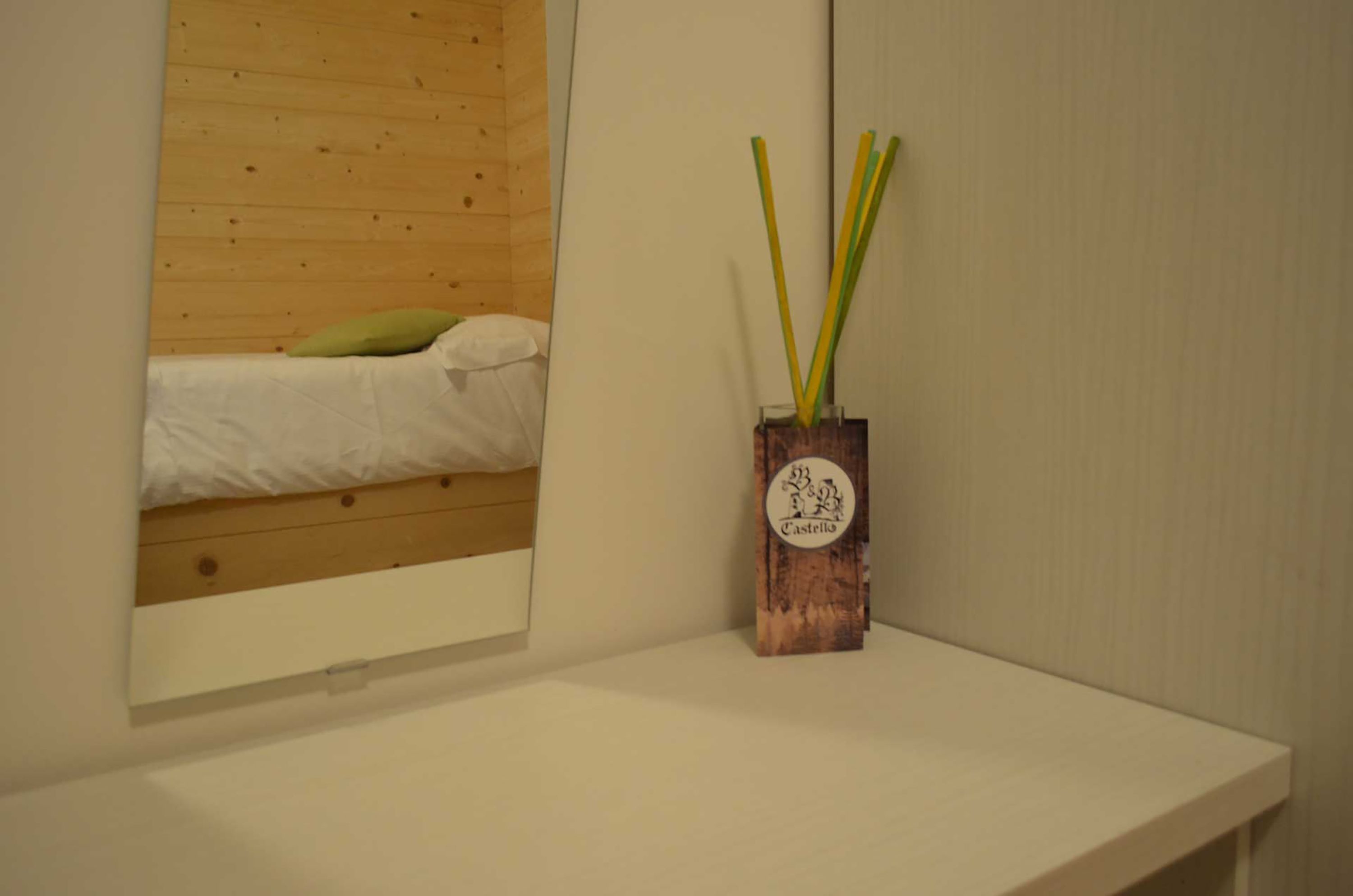 Un lit en bois avec un oreiller vert et une couverture blanche, un vase en bois contenant des branches vertes et jaunes, et une étagère blanche.
