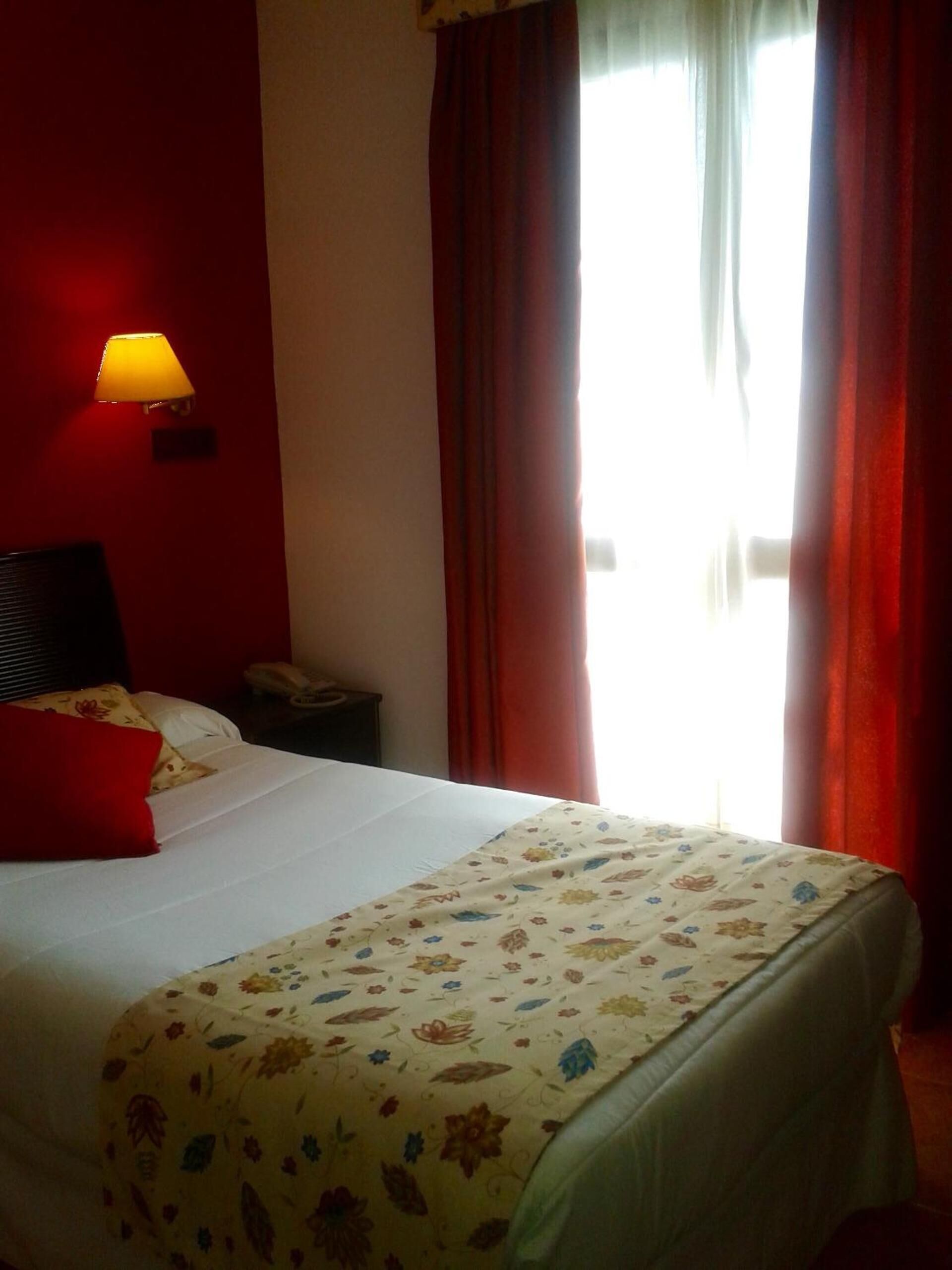 Una habitación de hotel con cama, lámpara de noche, teléfono y ventana con cortinas.