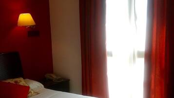 Una habitación de hotel con cama, lámpara de noche, teléfono y ventana con cortinas.