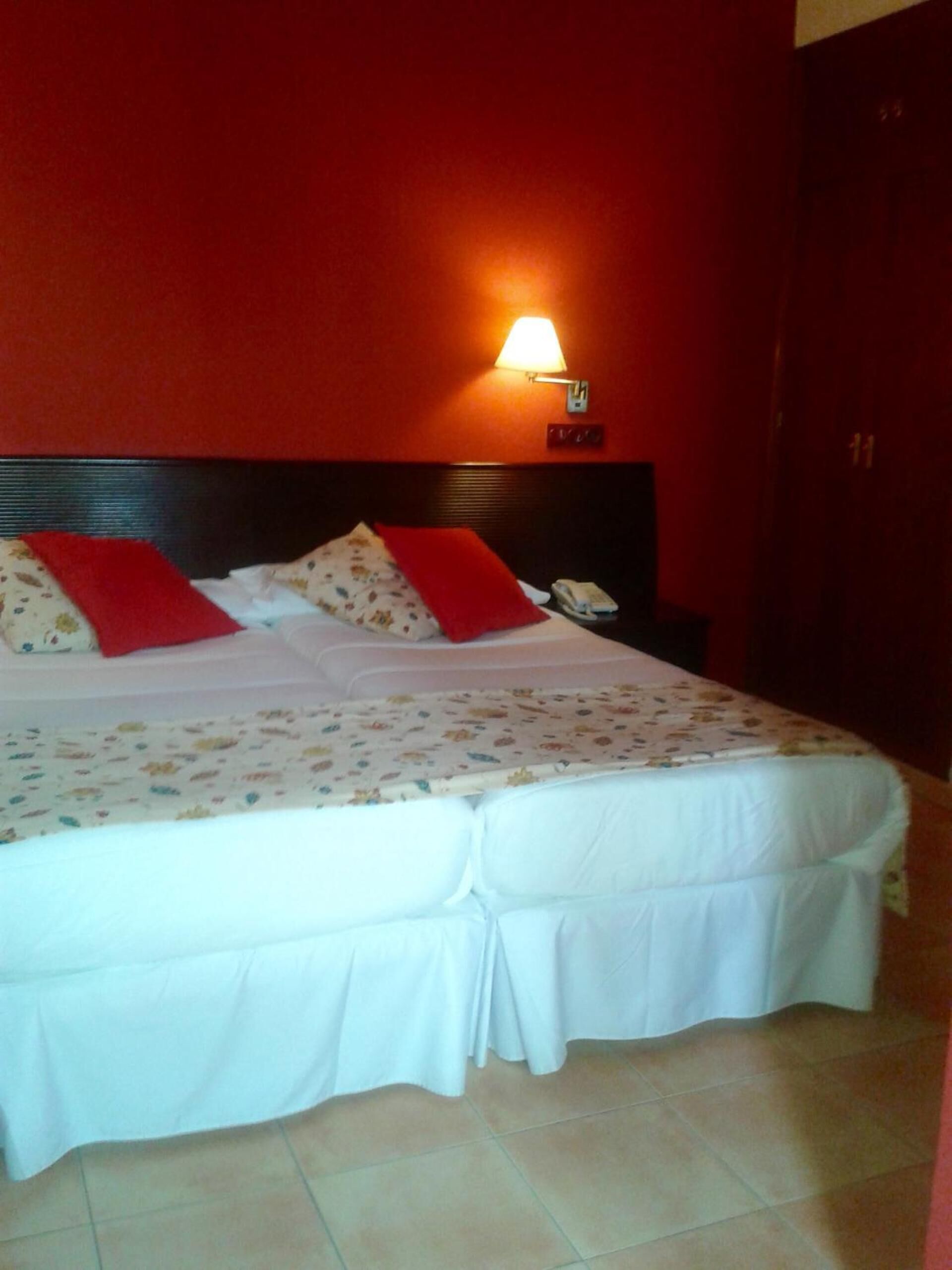 Cama doble con ropa de cama blanca y almohadas rojas, una mesita de noche con lámpara y una pared roja.
