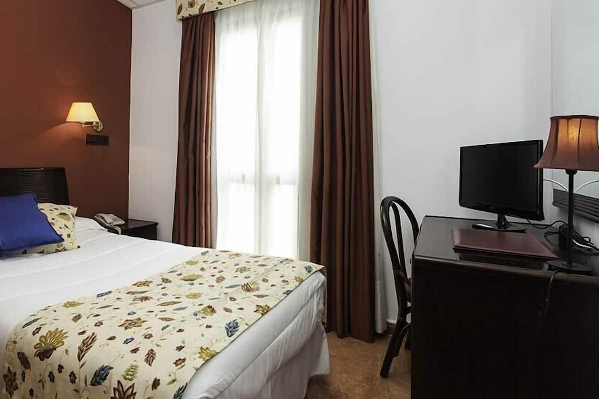 Ein Hotelzimmer mit einem Bett, einem Schreibtisch mit Computer, einer Lampe und einem Fenster mit Vorhängen.