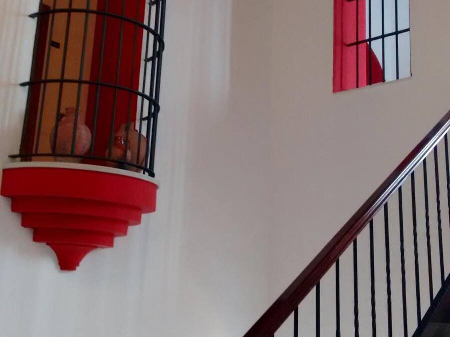 Una escalera con barandilla de madera y balaustres negros, que conduce a un rellano con una ventana de marco rojo y barandilla de balcón también roja.
