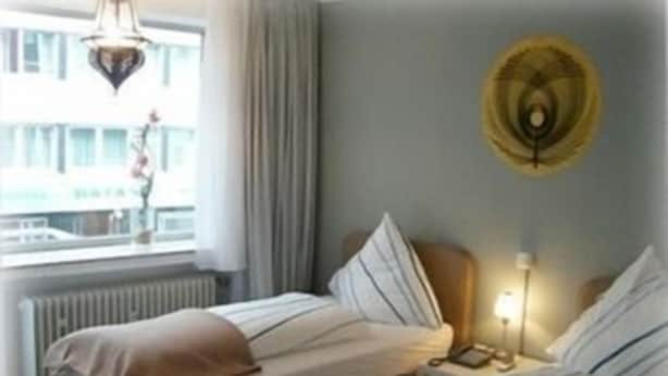 A hotel room with two beds, a window with curtains, a radiator, and a wall clock. - CityMinis (Düsseldorf)