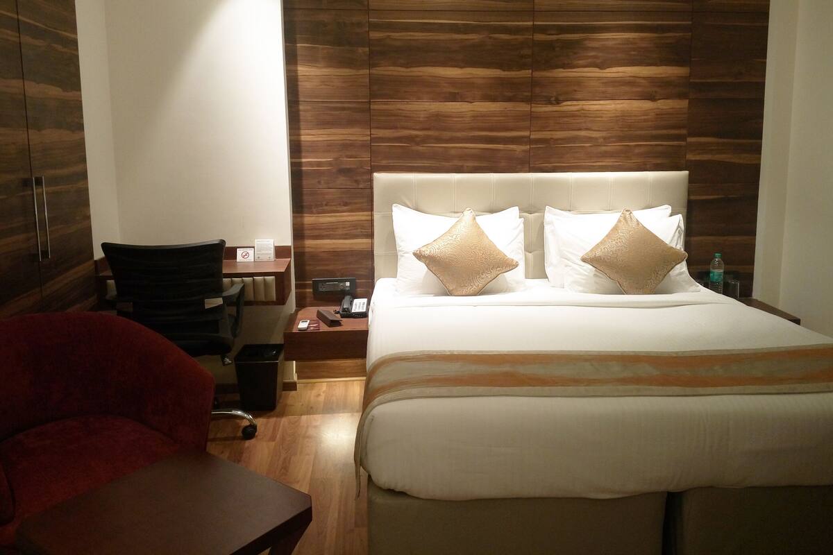 A hotel room with a wooden panel wall, a bed with two decorative pillows, a desk with a chair, and a red armchair.