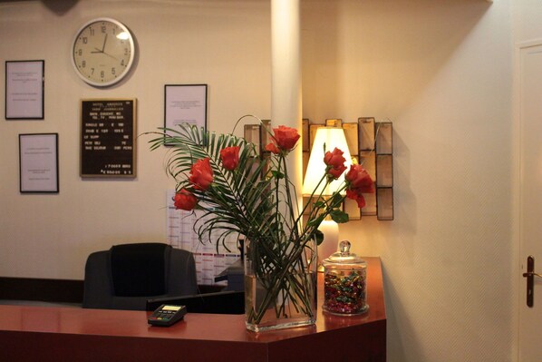 A reception desk with a clock, framed certificates, a vase of red roses, a lamp, and a jar of colorful candies. - Amadeus Paris (Paris)