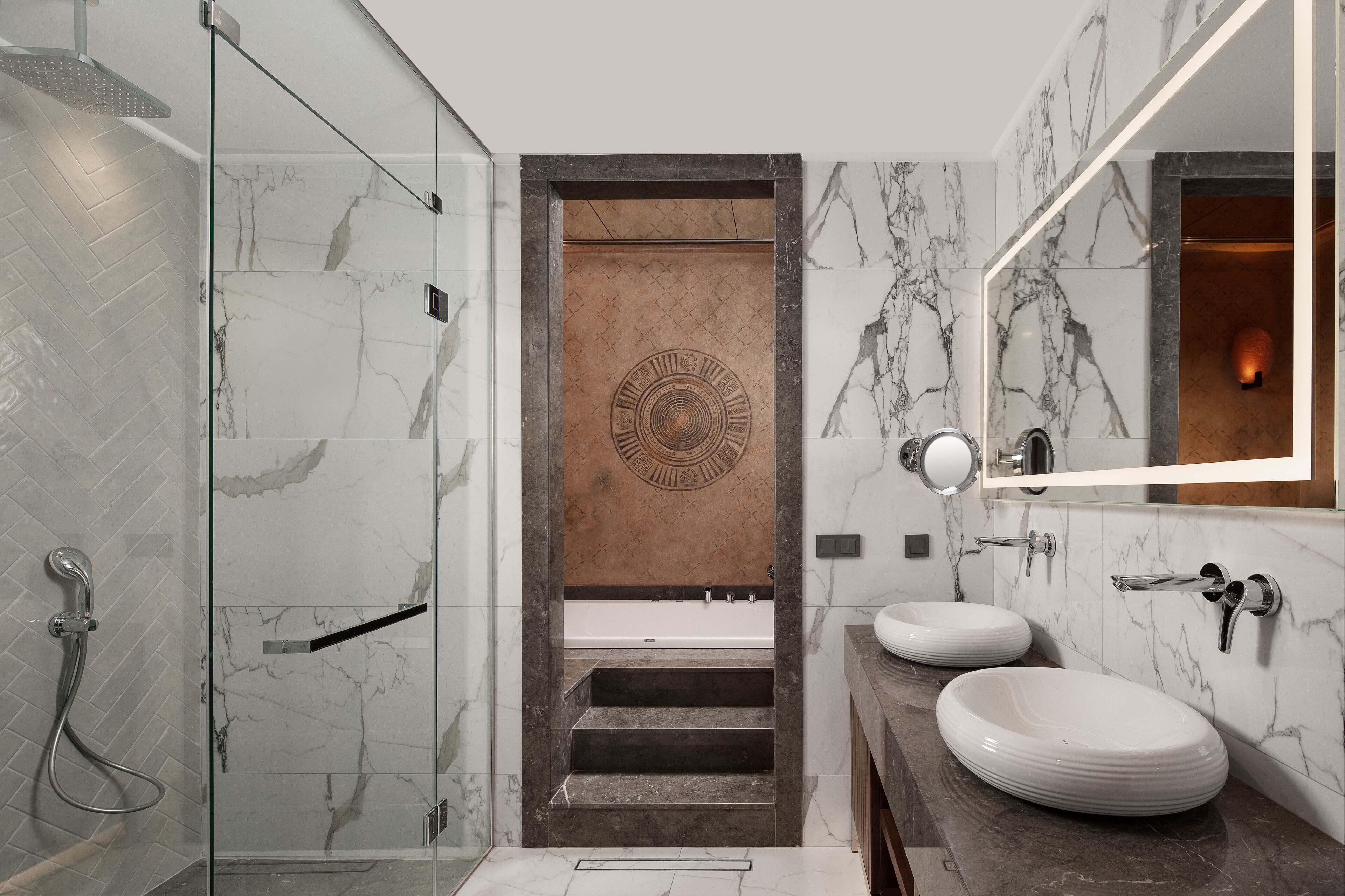A modern bathroom with a glass-enclosed shower, a wooden door with a circular design, and two white sinks with a marble backsplash.
