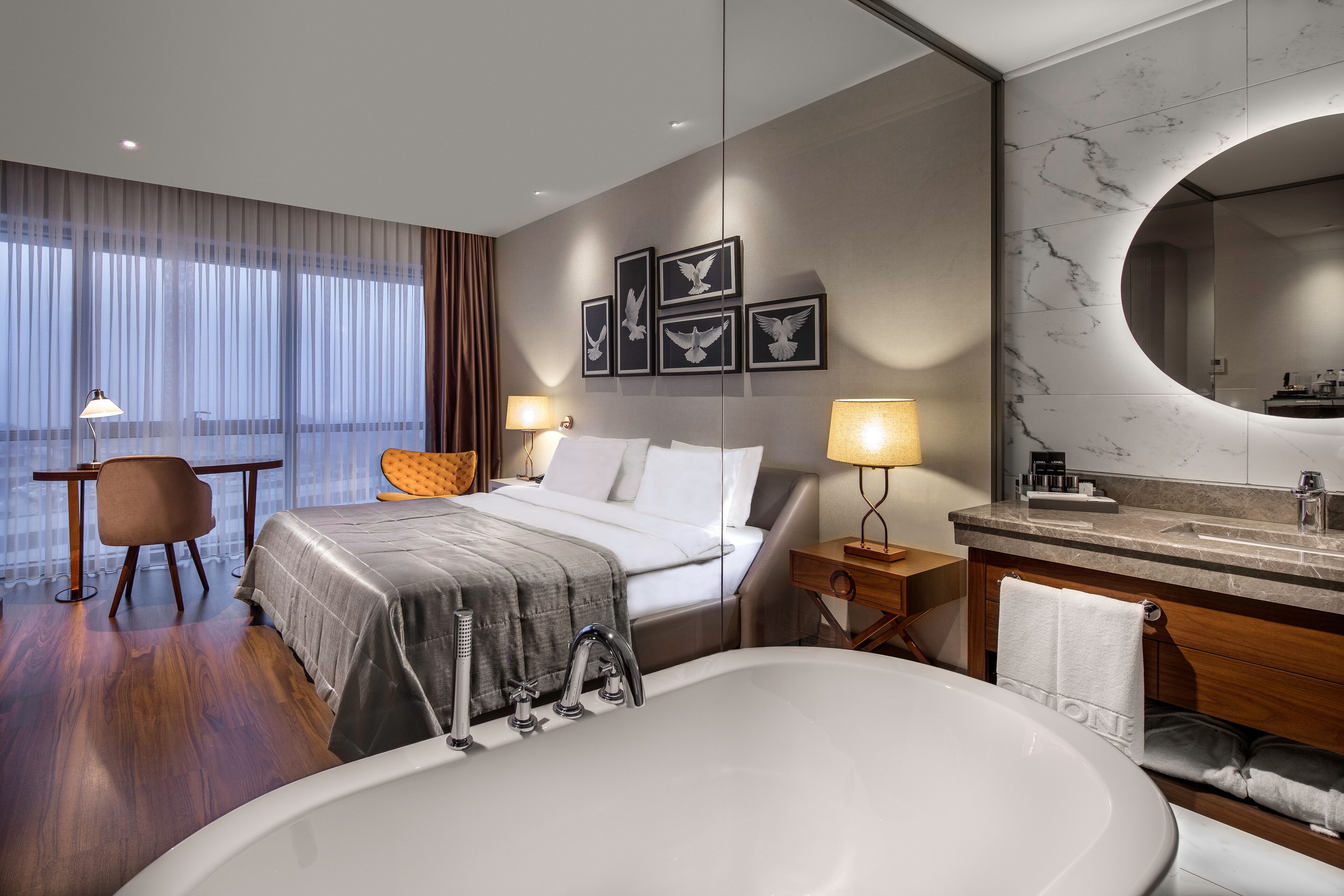 A modern hotel room with a large bed, a sitting area, a bathroom with a large mirror, and artwork on the wall.