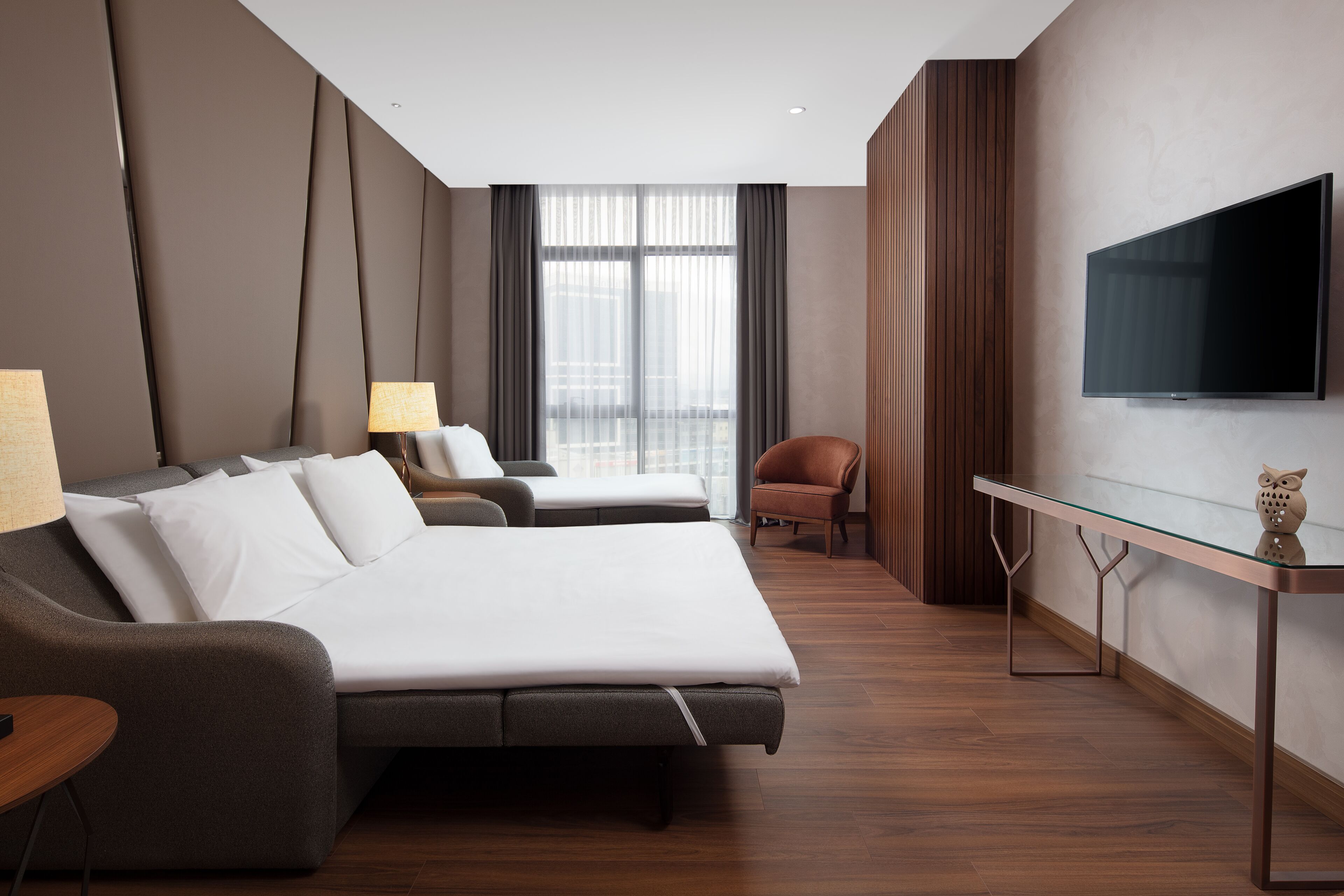 A modern hotel room with a large bed, a smaller sofa, a desk with a flat-screen TV, and a city view through large windows.