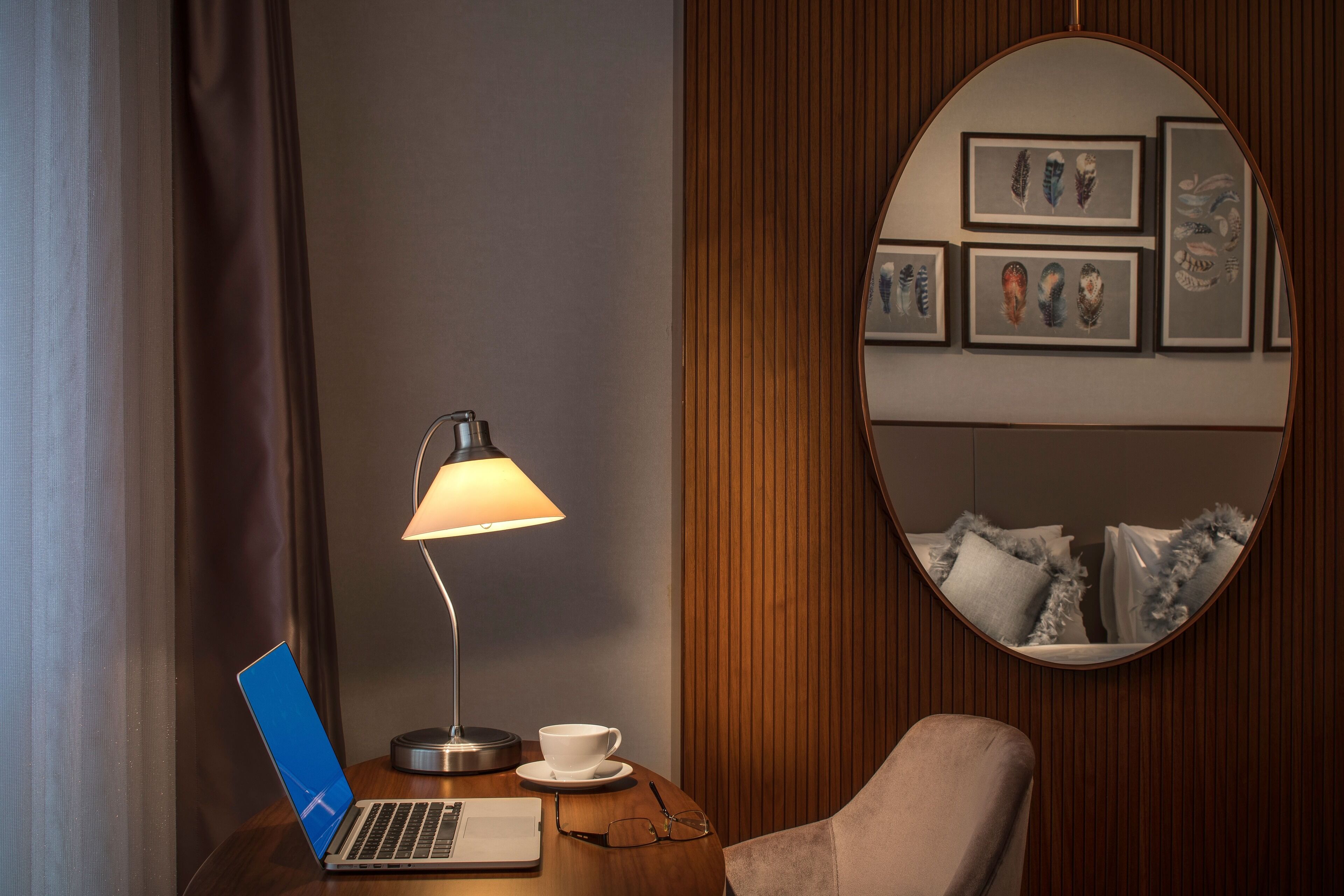 A room with a laptop on a wooden table, a lamp, a cup, and a chair. There is a mirror on the wall reflecting a bed with pillows.