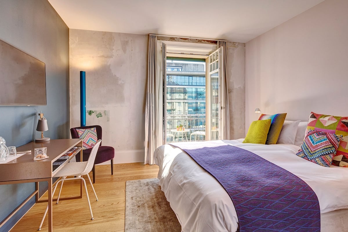 A modern bedroom with a large bed, a desk, and a chair. There is a window with a view of buildings outside.