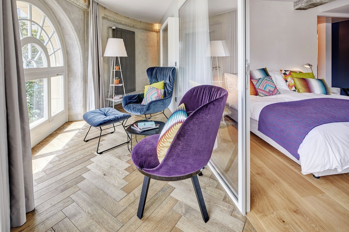 A modern hotel room with a large bed, two armchairs, a small table, and a large window.