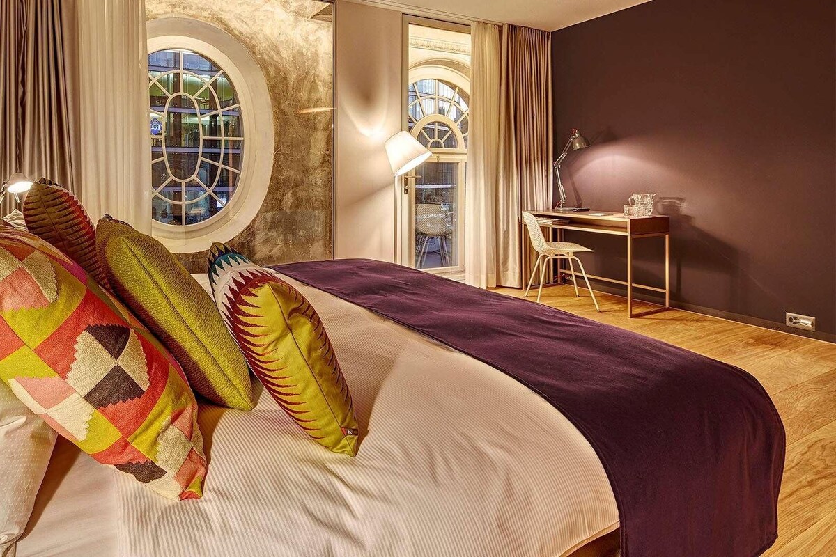 A bedroom with a bed, a desk, and two windows with curtains.