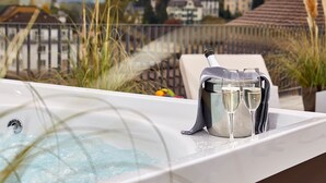 Un jacuzzi con una botella de champán y copas, rodeado de plantas y con vistas panorámicas a casas y montañas.